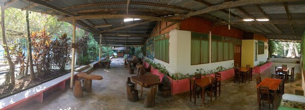 Un espace de restauration en plein air, avec des tables et des bancs en bois, entouré de plantes en pot et d’arbres.
