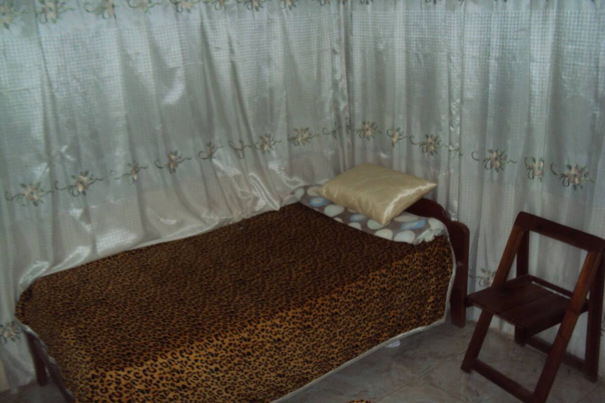 Una habitación pequeña con cama individual, colcha de estampado de leopardo, una silla de madera y una ventana con cortinas de gasa.