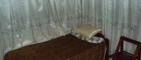 Een kleine eenpersoonskamer met een luipaardprint sprei, een houten stoel en een raam met dunne gordijnen.