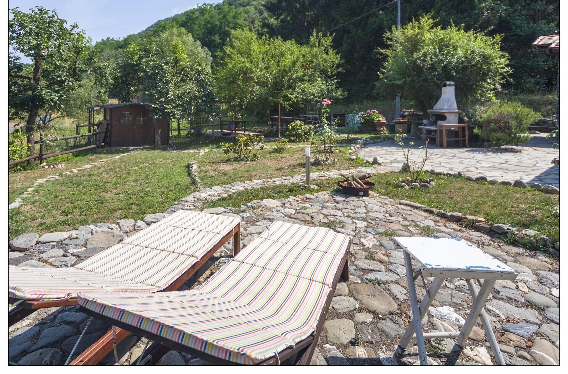 A rustic outdoor seating area with striped lounge chairs and a stone table, surrounded by a stone pathway and lush greenery.