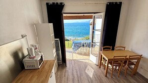 Ett kompakt kök med mikrovågsugn, kaffebryggare och ett litet kylskåp. Det finns ett matbord i trä med stolar och en blå säng. En balkong med utsikt över havet syns genom en öppen dörr.