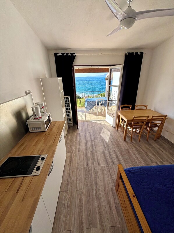 A compact kitchen with a microwave, coffee maker, and a small refrigerator. There is a wooden dining table with chairs and a blue bed. A balcony with a view of the sea is visible through an open door.