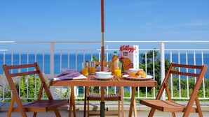 A table set with breakfast items, including cereal and juice, on a balcony with a view of the ocean.