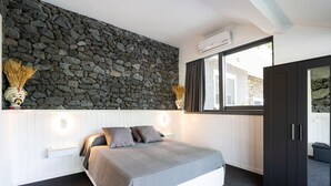 Une chambre avec un mur en pierre, un lit avec une literie grise, une table de chevet et une armoire.