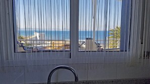 Ein Zimmer mit Meerblick, durchscheinende Vorhänge, ein Waschbecken und ein Fenster mit Blick auf Liegestühle und Sonnenschirme.