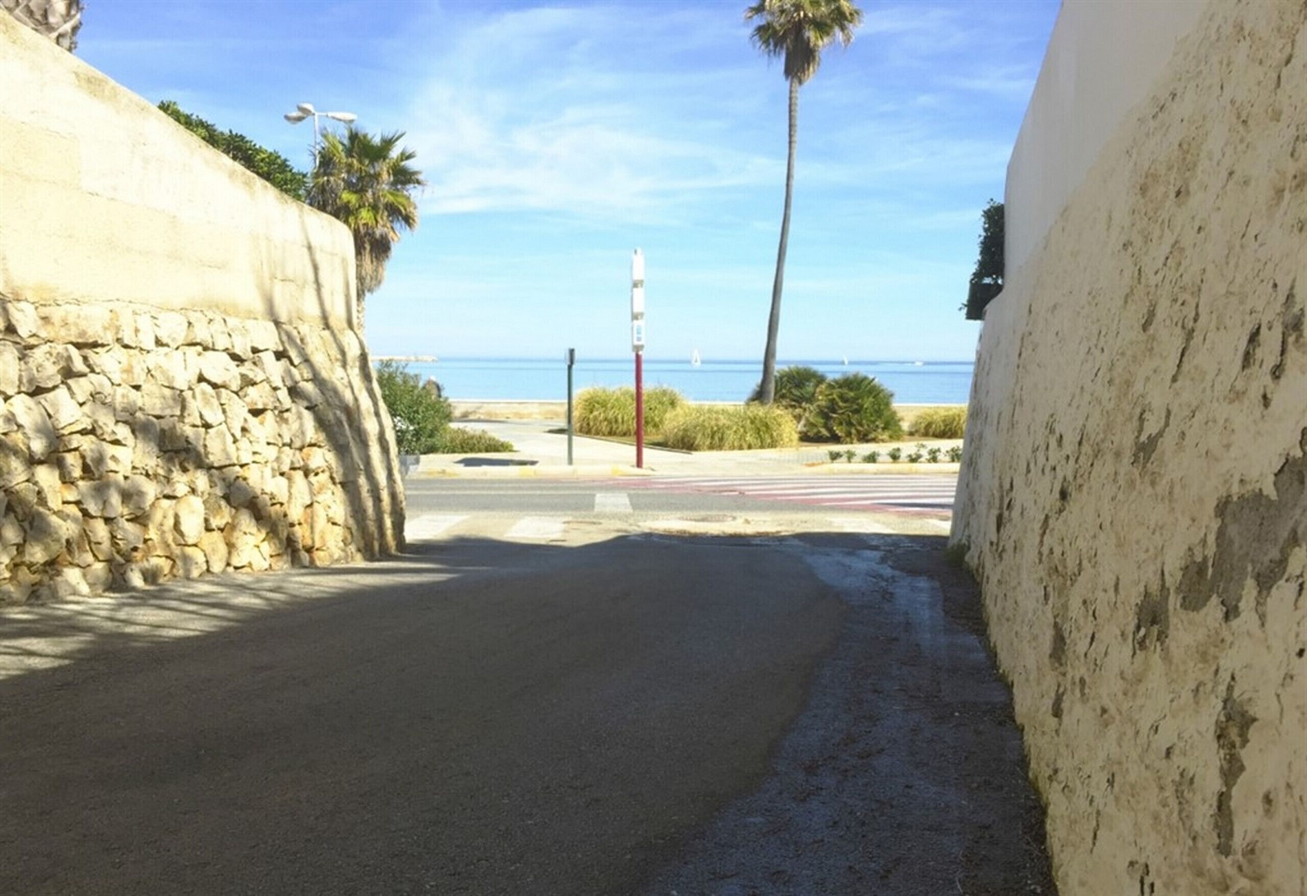 A narrow street with a stone wall on one side and a white wall on the other, leading to a beach with palm trees and a clear sky.