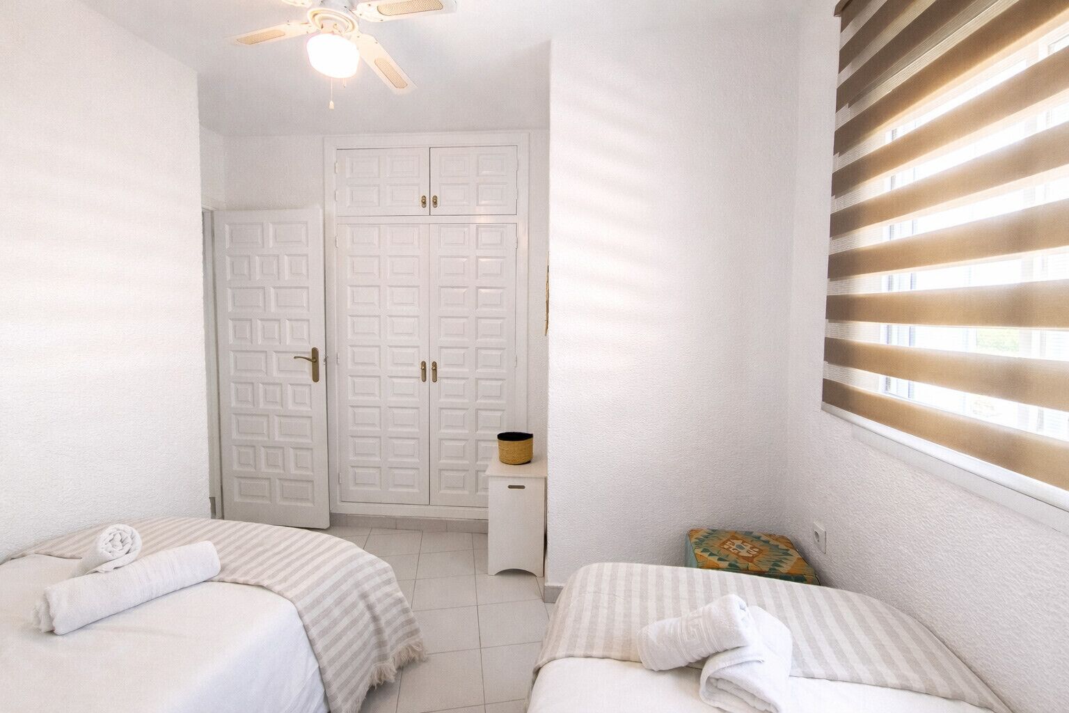 A bedroom with two beds, a wardrobe, a ceiling fan, and a window with blinds.