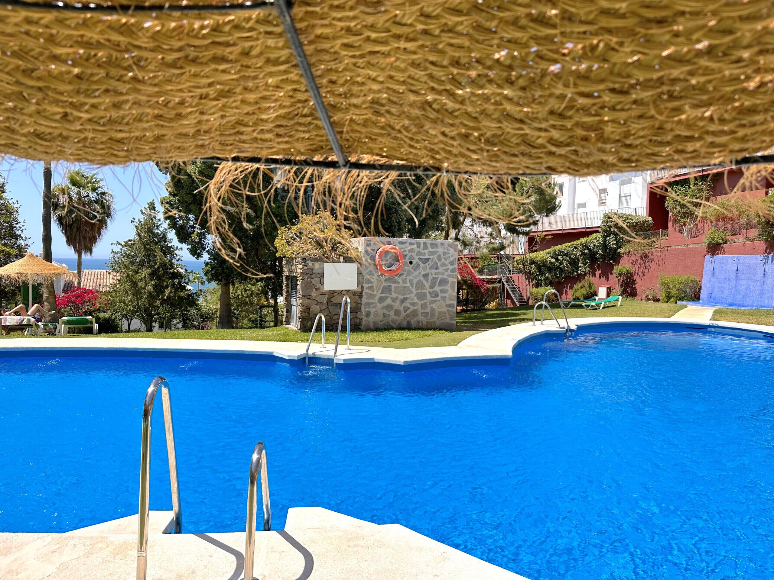 A swimming pool with a stone wall and a lifebuoy, surrounded by greenery and a building in the background.