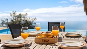 A dining table set with plates, cutlery, a basket of bananas, and glasses of orange juice, overlooking a pool and ocean view. - 17°West – Holiday villa (option with heated pool) (Estreito da Calheta)