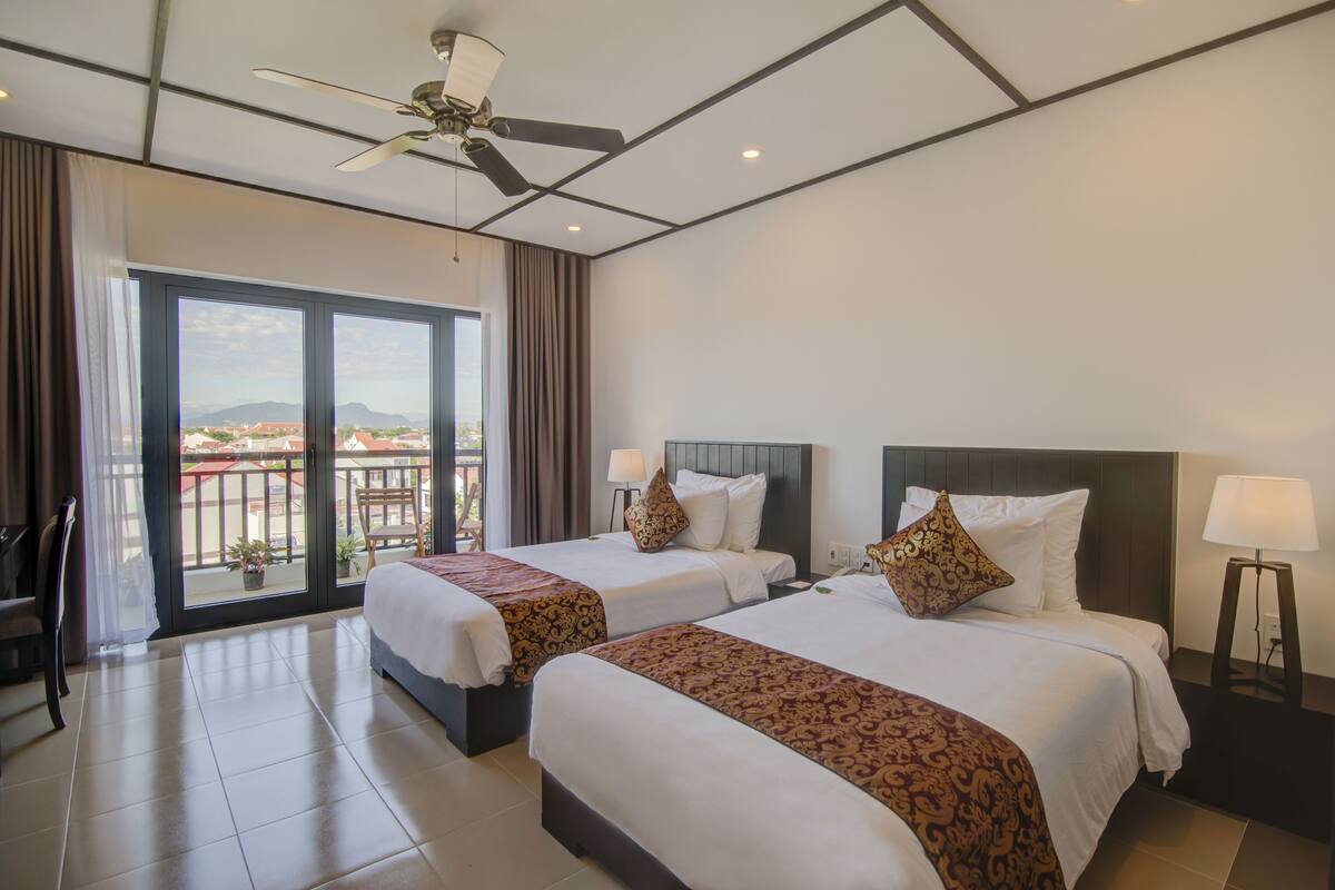 A hotel room with two beds, a ceiling fan, a balcony with a view, and a nightstand with a lamp.