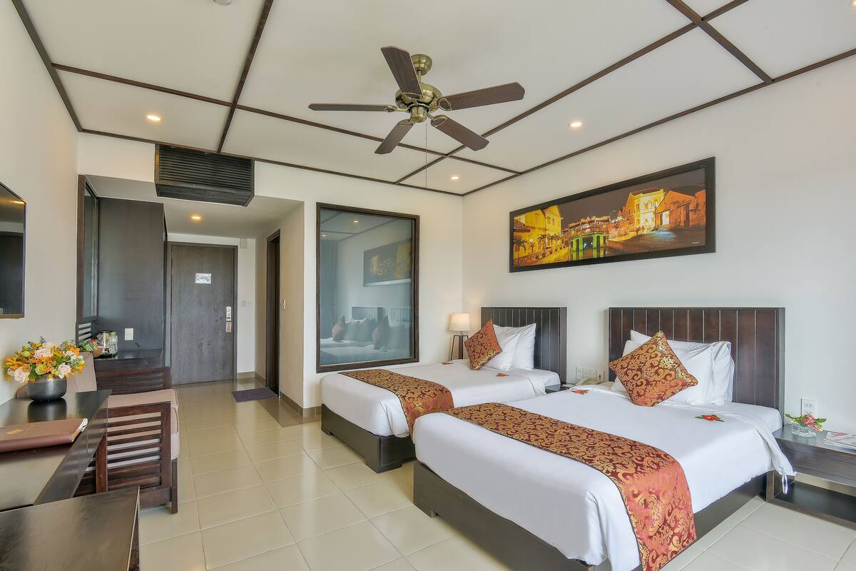 A hotel room with two beds, a ceiling fan, a painting, and a dining area with a table and chairs.
