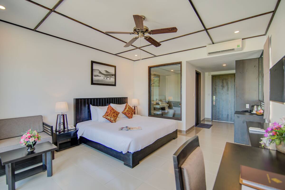 A hotel room with a bed, bedside tables, a desk, a chair, a ceiling fan, and a bathroom visible through a glass door.