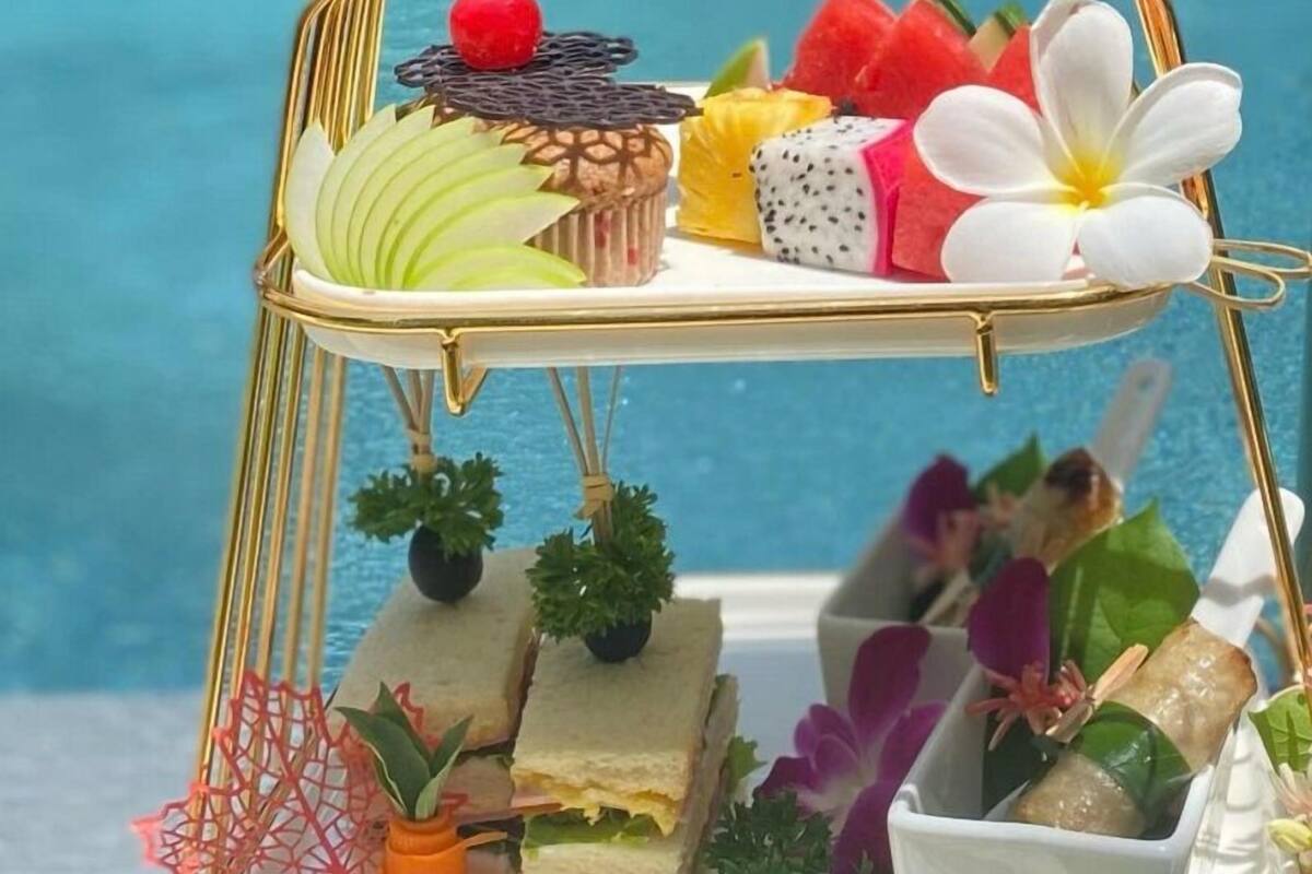 A tiered dessert tray with various sweets and fruits, a small plant, and a decorative item with a golden top, placed on a table near a pool.