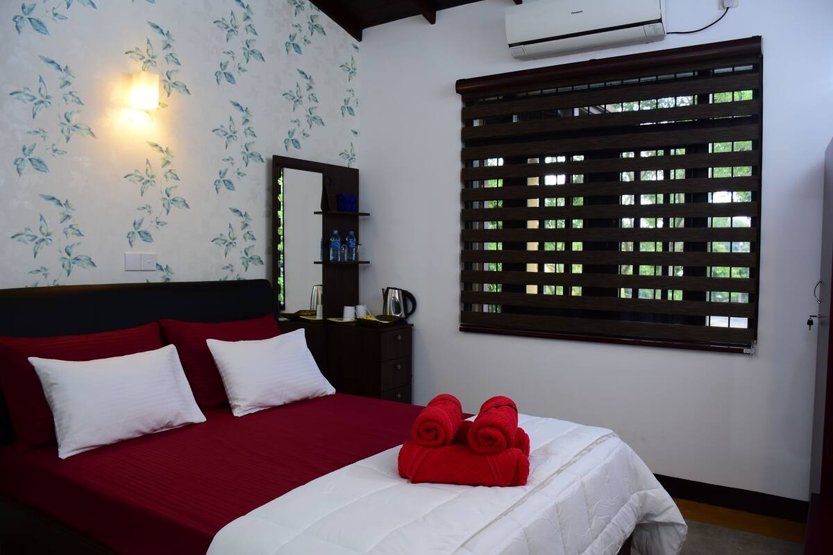 A hotel room with a bed, red and white bedding, a nightstand with a mirror, a wall-mounted air conditioner, and a window with a wooden blind.