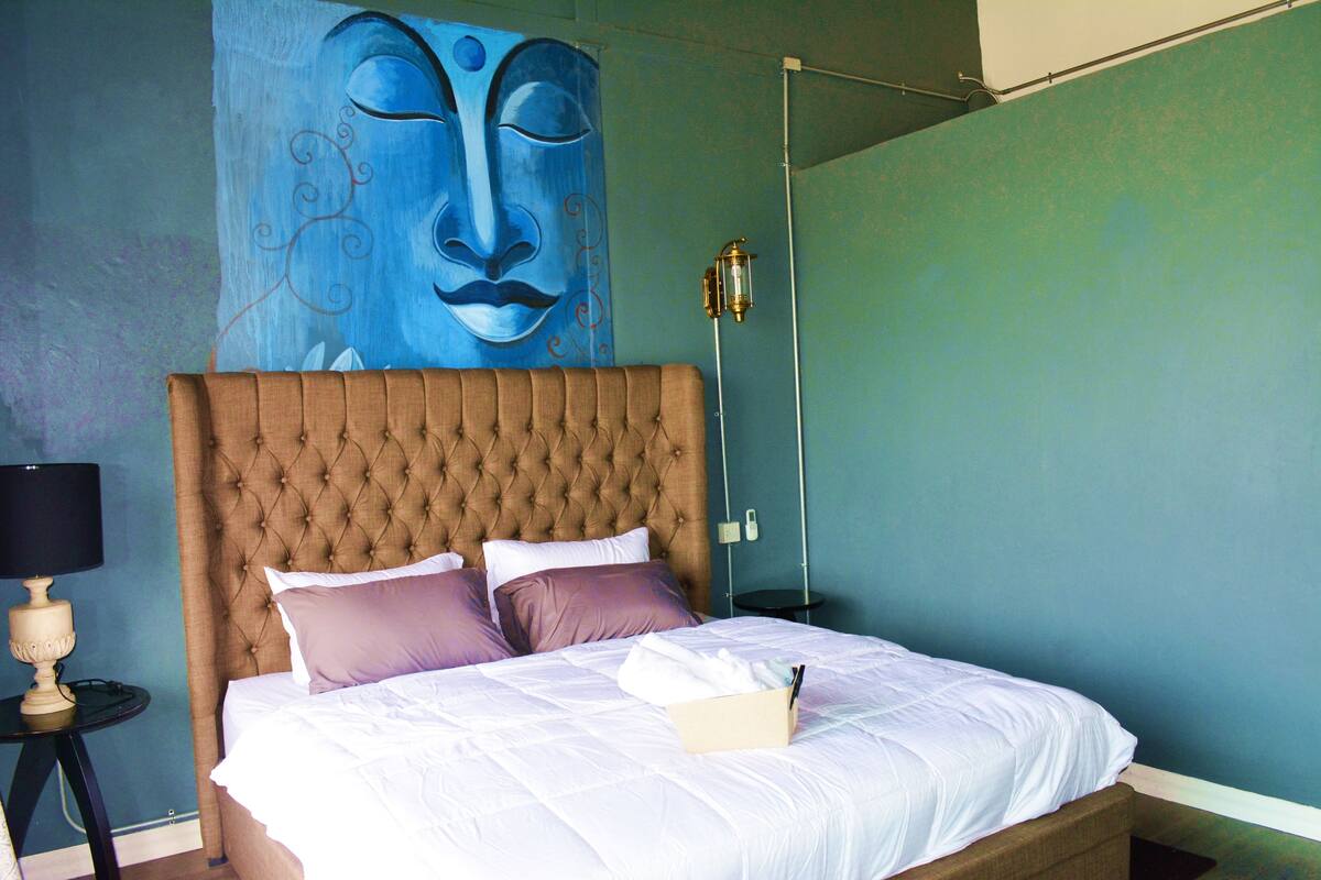 A bedroom with a tufted headboard, a bed with white and purple bedding, a nightstand with a lamp, and a wall mural.