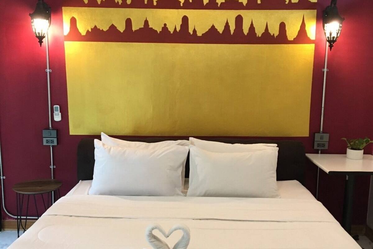 A hotel room with a large bed, white bedding, a heart-shaped towel arrangement, and a yellow wall with a silhouette of a city skyline.