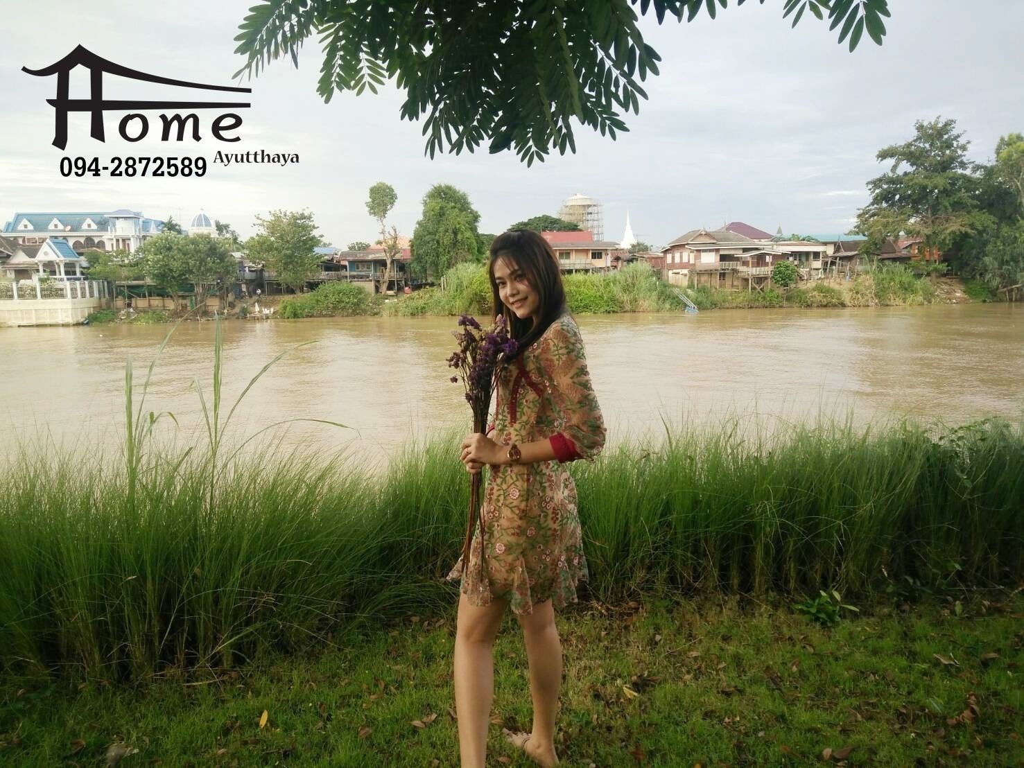 A person standing by a river with a house and buildings in the background.