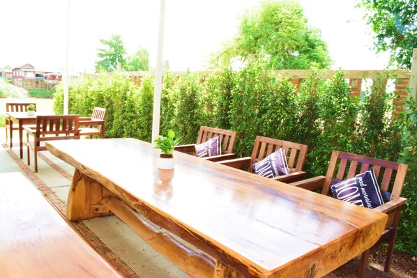 Un espacio exterior para comer con una larga mesa de madera y varias sillas de madera, rodeado de abundante vegetación.
