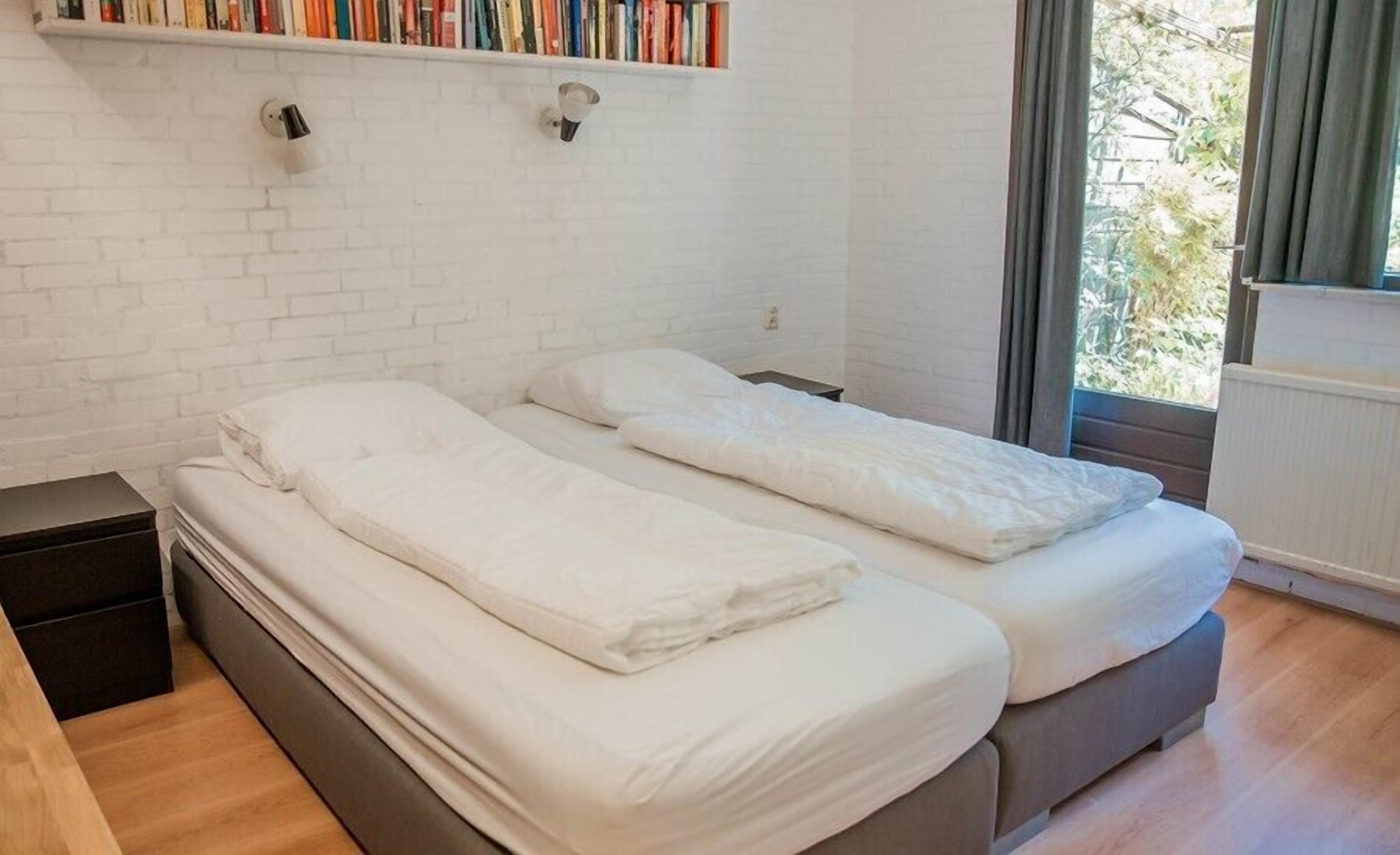 A bedroom with two single beds, a white brick wall, a bookshelf with books, and a window with curtains.
