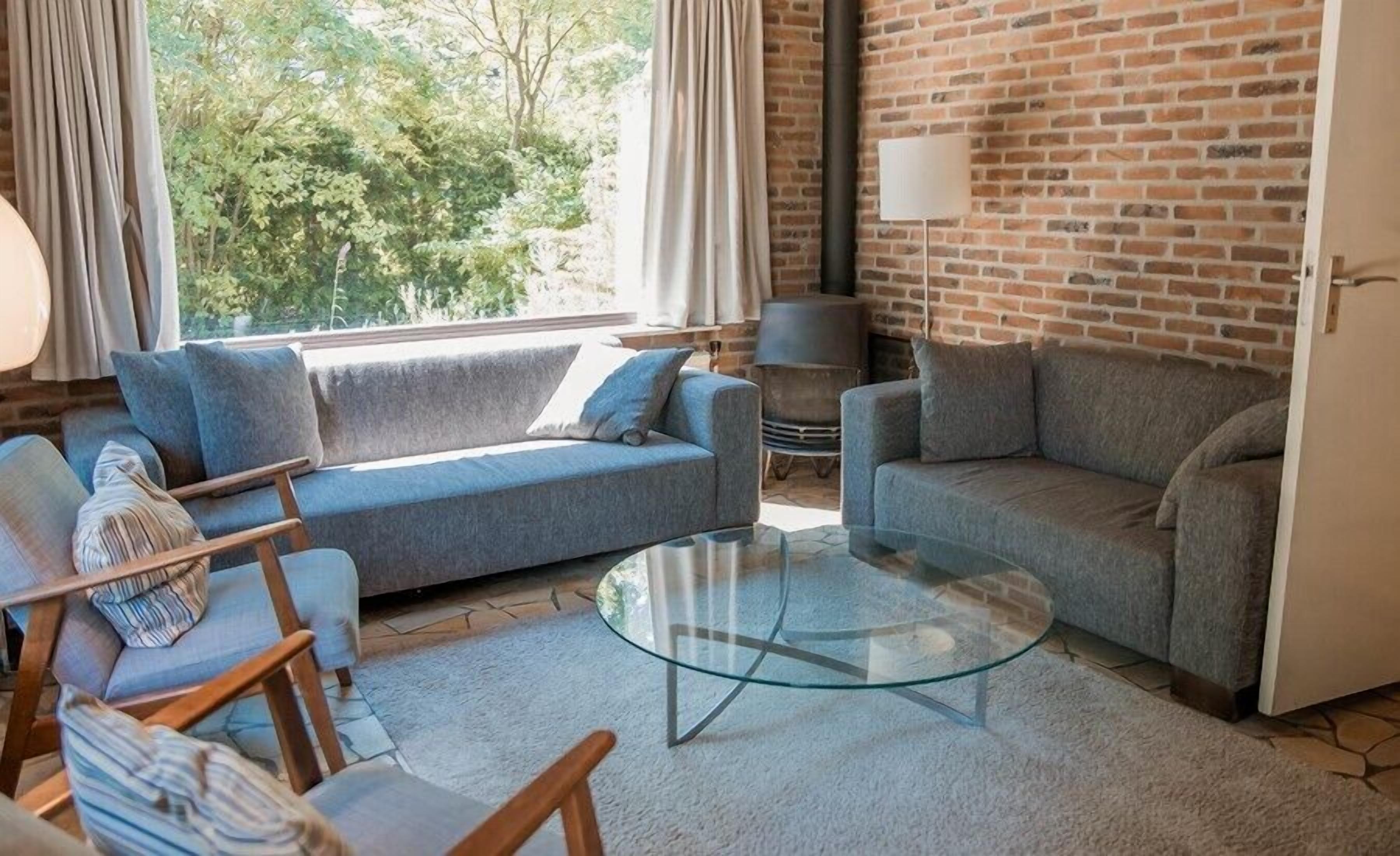 A living room with a brick wall, two grey sofas, a glass coffee table, and a view of trees through large windows.