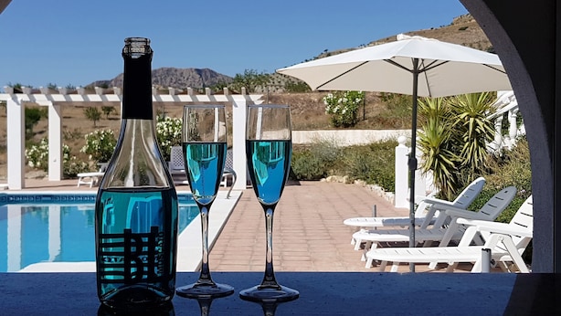 A poolside setting with a bottle and two glasses of blue liquid, white lounge chairs, and a white umbrella.