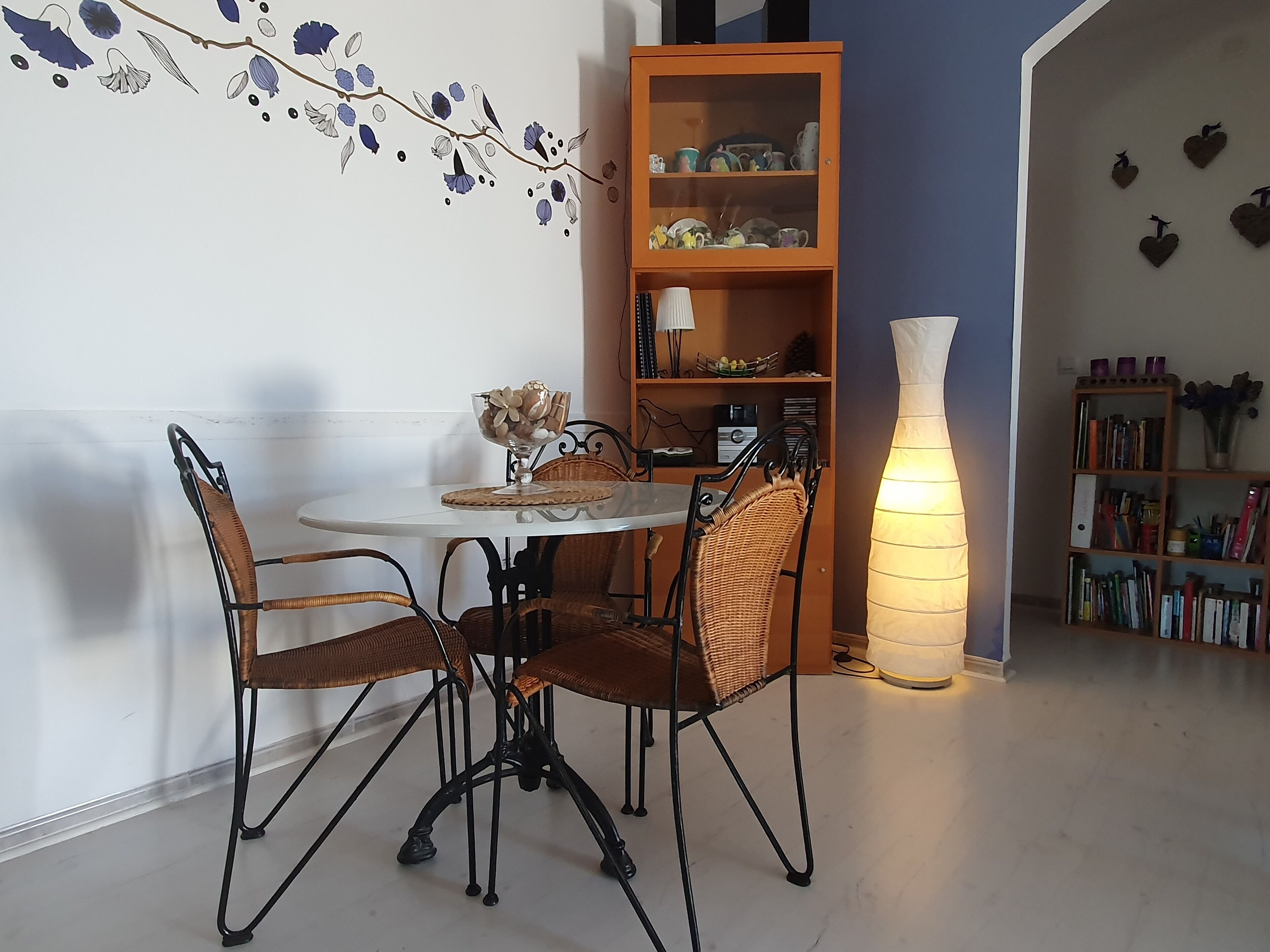 Un comedor con una mesa redonda, cuatro sillas de mimbre, un florero con flores y una estantería llena de libros y adornos.