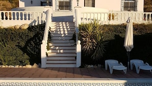 A white building with a tiled roof, a staircase leading to a balcony, and a pool in the foreground.