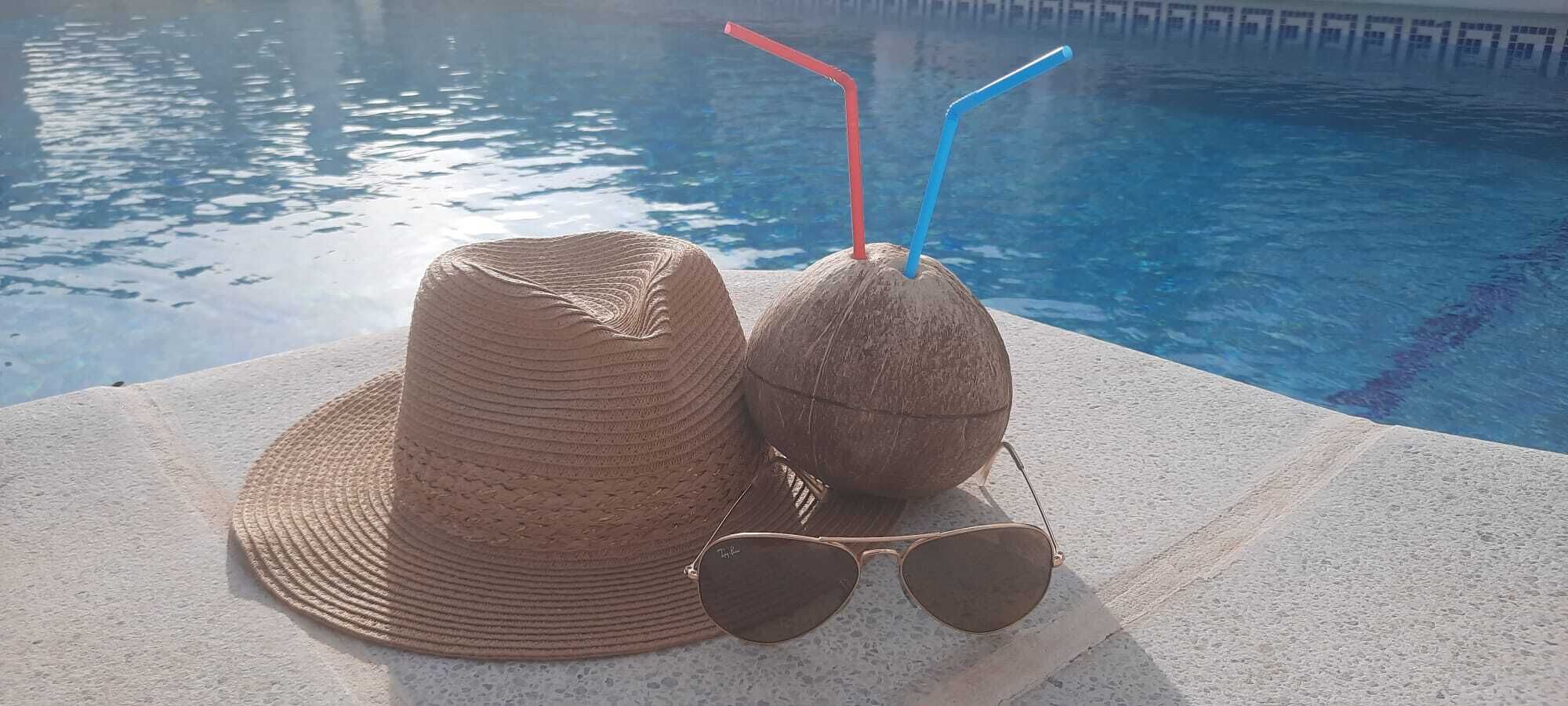A straw hat, sunglasses, and a coconut with straws by a pool.