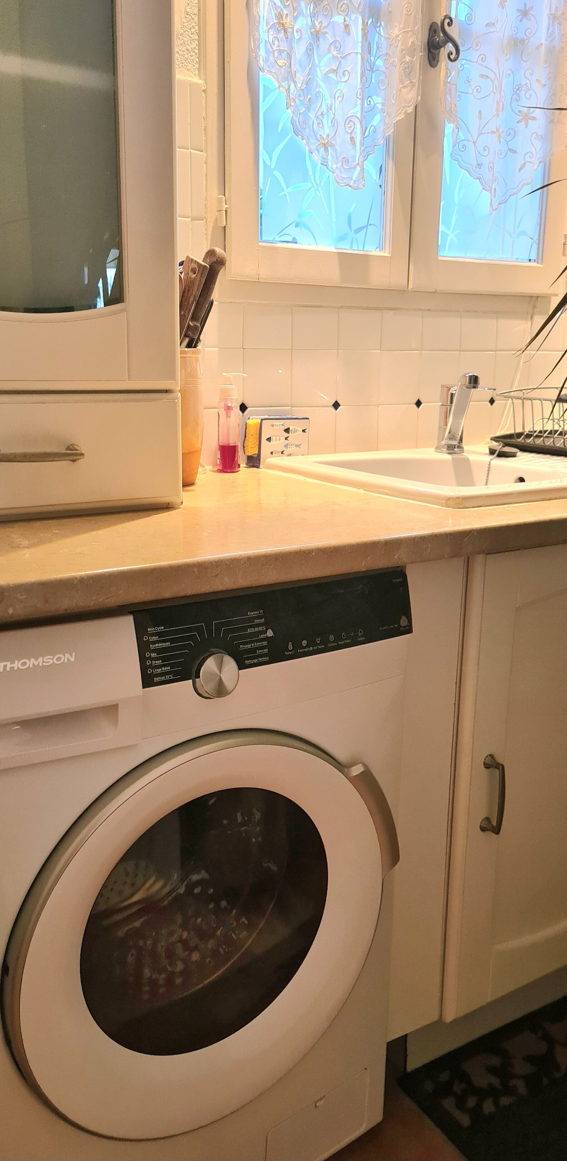 A compact kitchen with a built-in dishwasher, a washing machine, a sink, and a window with decorative curtains.