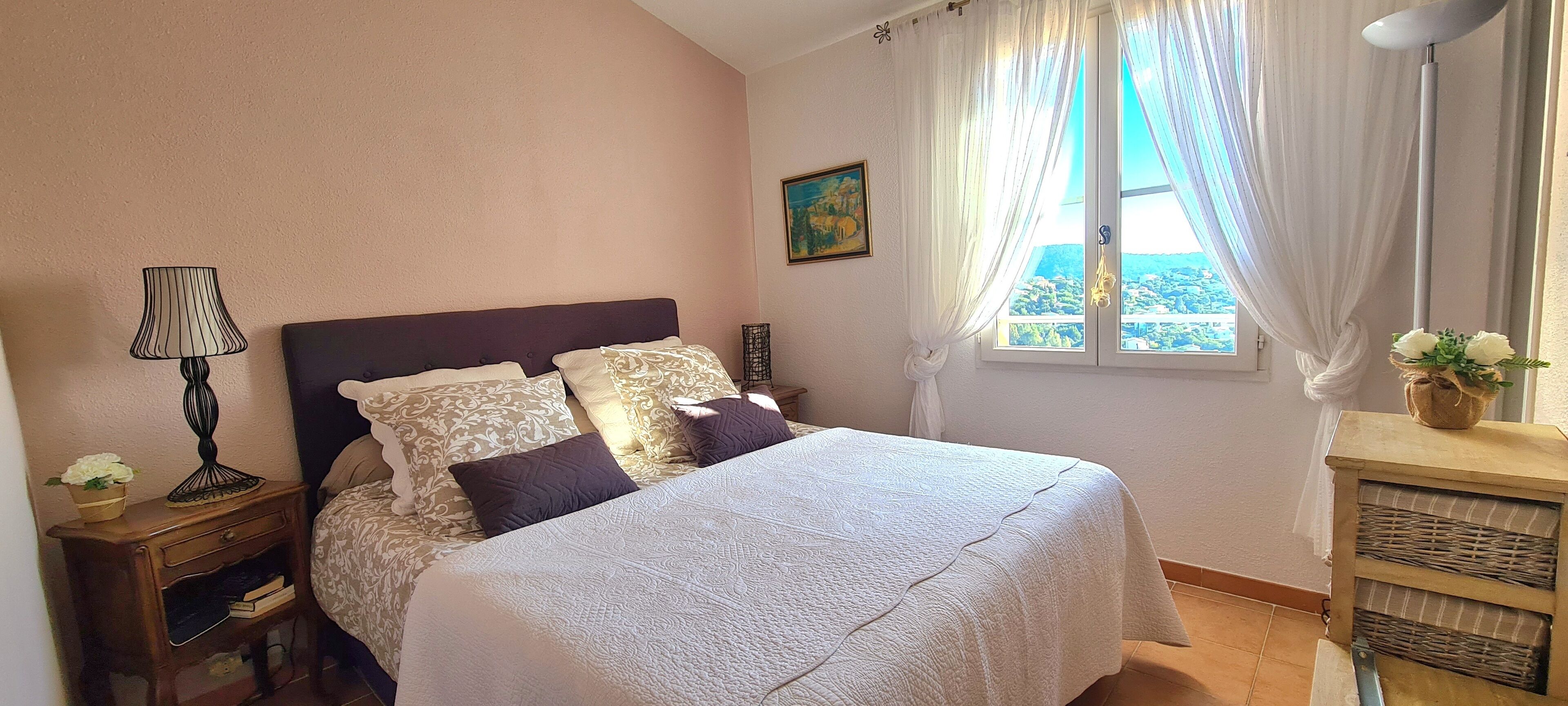 Quarto com cama, mesa de cabeceira, luminária, janela com cortinas, quadro e vista para o exterior.
