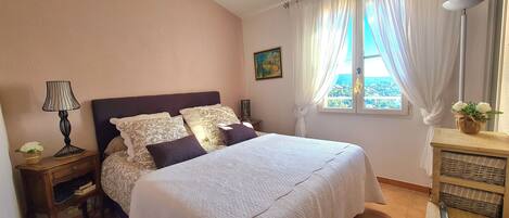 Quarto com cama, mesa de cabeceira, luminária, janela com cortinas, quadro e vista para o exterior.