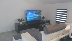 A modern living room with a flat-screen TV, a coffee table, a sofa, and a window with blinds. - Saint Aubin de Médoc will take place from August 22 to 29, 2026 (Saint-Aubin-De-Médoc)