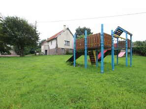 A playground with a slide and climbing structure in a grassy field, with a house in the background.