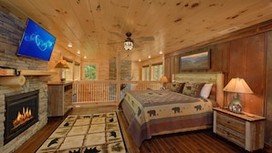 A log cabin bedroom with a fireplace, a bed with a bear-themed quilt, a television mounted on the wall, and a stone fireplace.