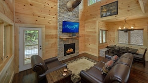 A log cabin interior with a stone fireplace, leather seating, a television, and a dining area.