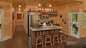 A wooden cabin interior with a kitchen featuring a refrigerator, stove, and microwave. There are wooden stools and a table with a light fixture above.