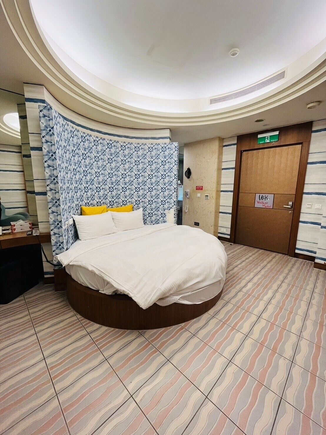 Habitación de hotel con una cama redonda, un cabecero estampado, una puerta de madera y un suelo de baldosas.