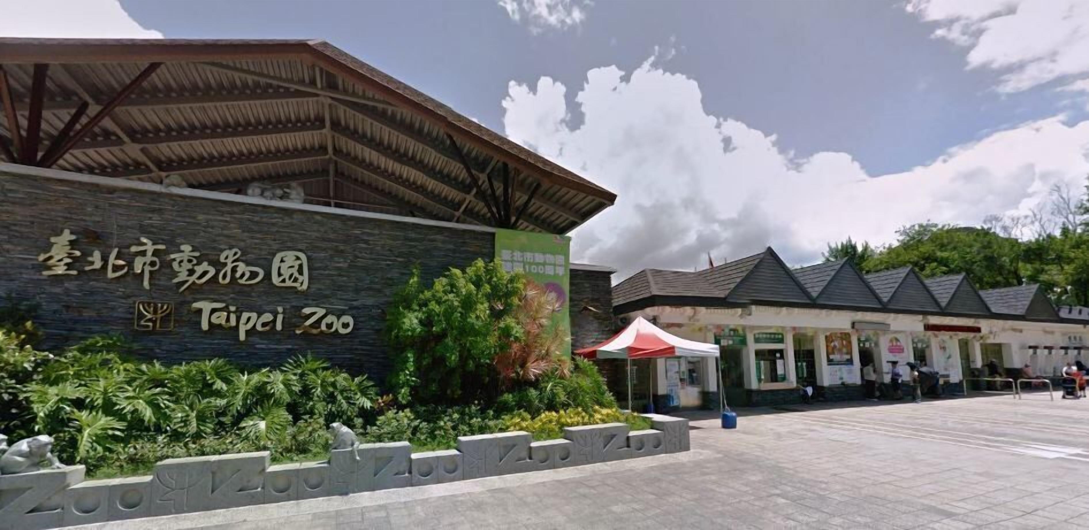 The entrance to Taipei Zoo with a stone wall and a sign, surrounded by greenery and a covered area with a red umbrella.