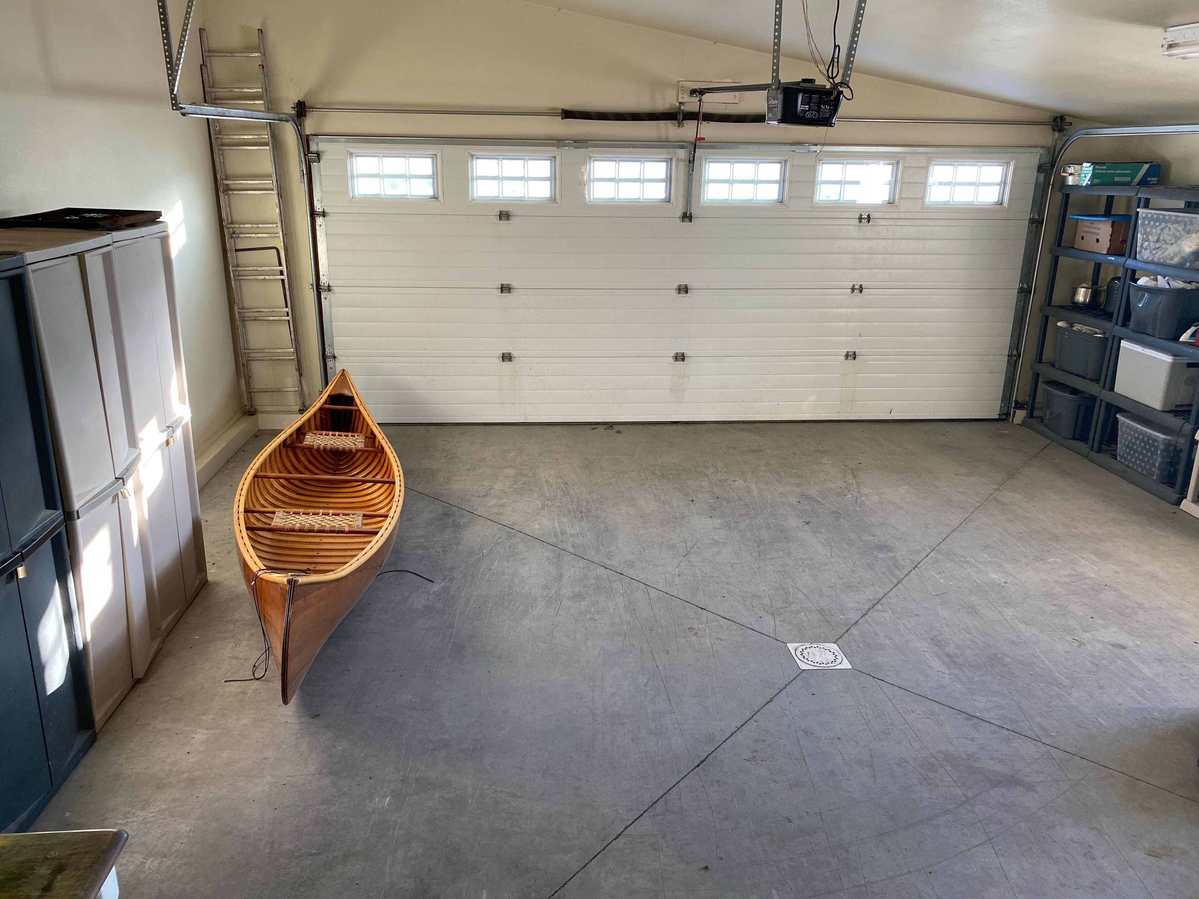 Un garage avec une porte de garage blanche, un bateau en bois, une échelle et des étagères avec des bacs de rangement.