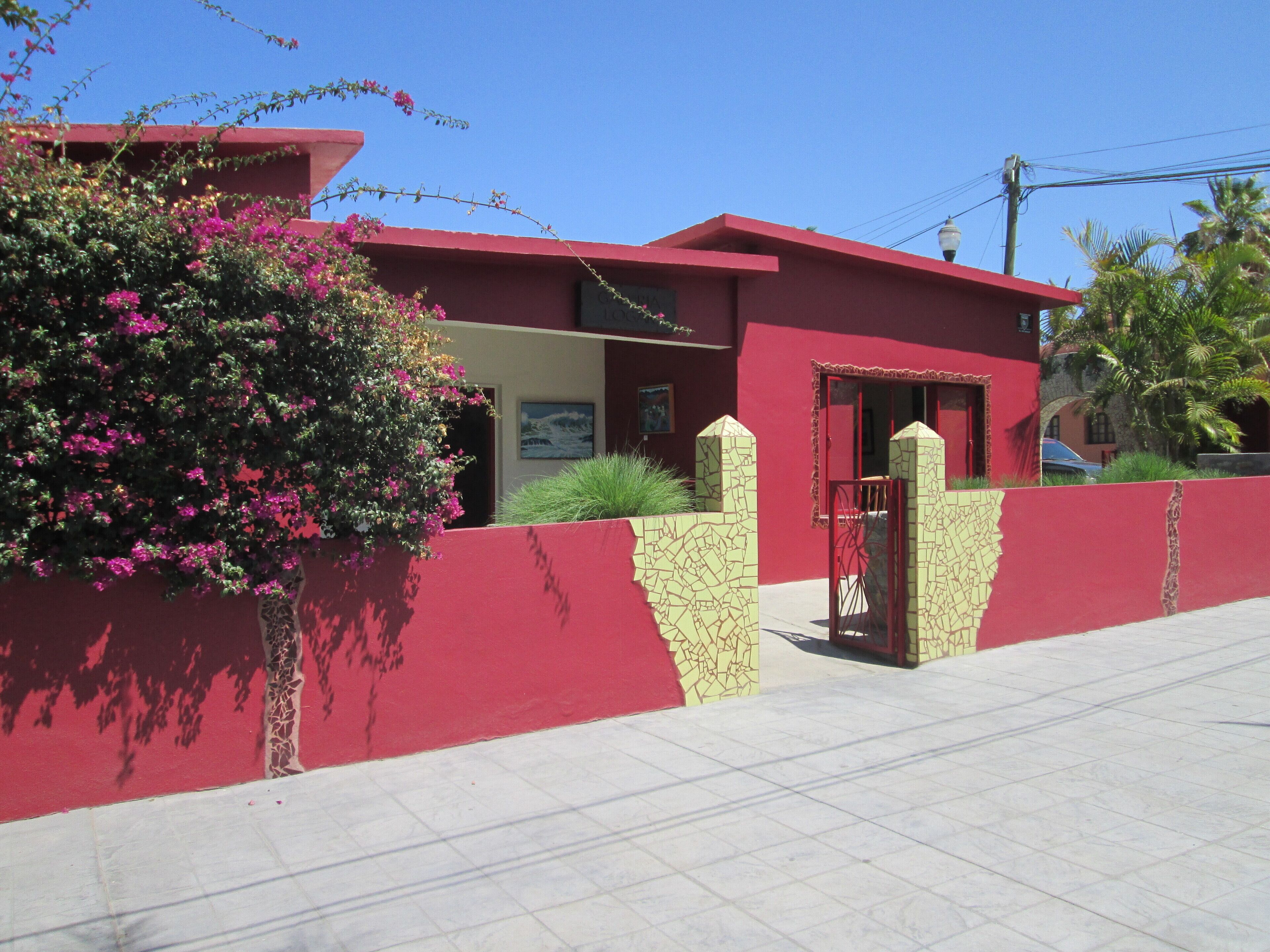 Un edificio rojo con entrada porticada, un pequeño jardín con flores rosadas y un cielo azul despejado.