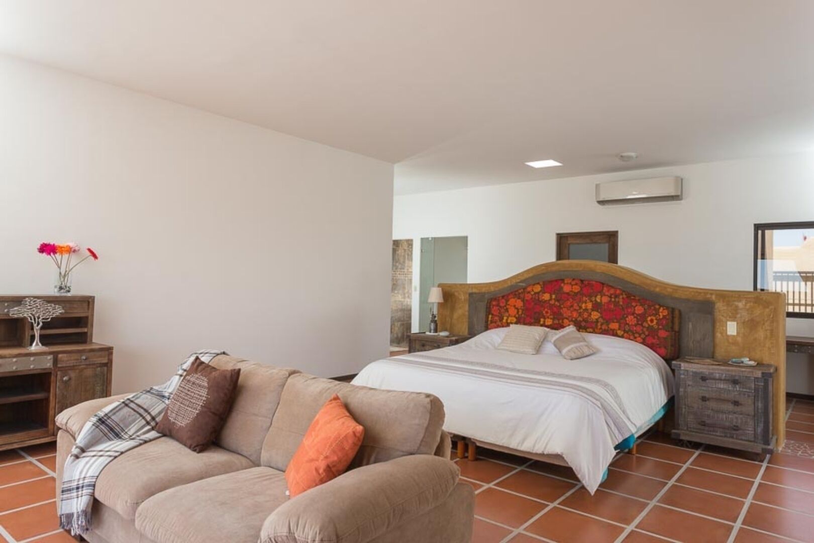 Une chambre à coucher avec un lit, une table de chevet en bois, une commode en bois, un canapé beige et une petite table sur laquelle est posé un vase de fleurs.