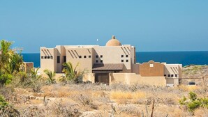 A desert-style building with a dome and multiple windows, surrounded by dry vegetation and overlooking a body of water. - Fabulous Fall and Winter:  HEATED POOL! (Todos Santos)