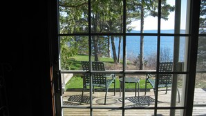 A view through a window with a grid pattern, overlooking a wooden deck with outdoor furniture and a body of water in the background.