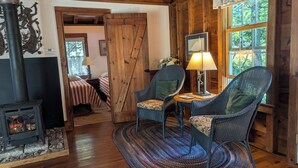 A cozy cabin interior with a wooden interior, a fireplace, two wicker chairs, a round table with a lamp, and a bed with striped bedding.