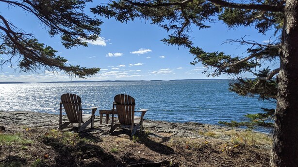 Two Adirondack chairs facing a lake with a rocky shoreline and trees framing the view.