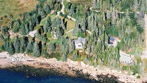 An aerial view of a coastal property with multiple houses surrounded by dense forests, a rocky shoreline, and a clear view of the sea.