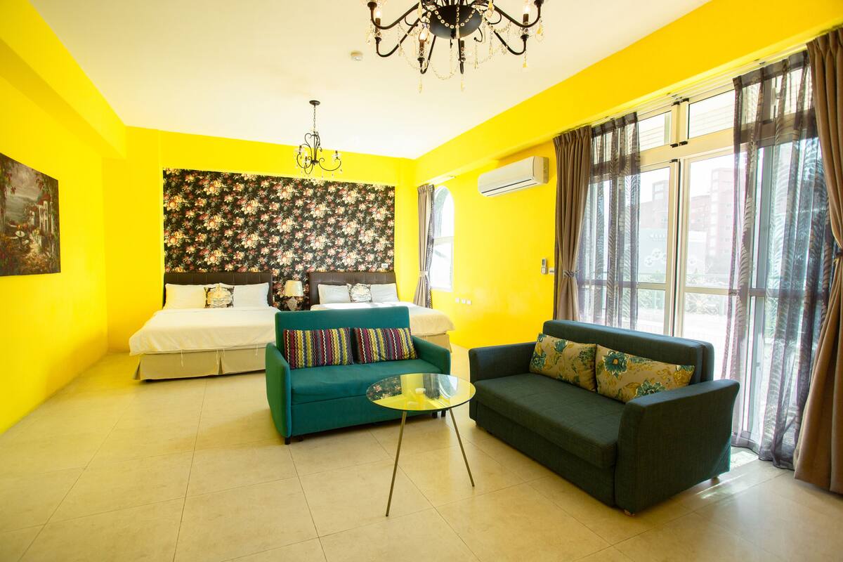 A bright yellow room with a bed, two sofas, a glass coffee table, and a floral-patterned headboard.