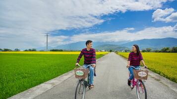 Two people riding bicycles on a paved path through a green field with mountains in the background.