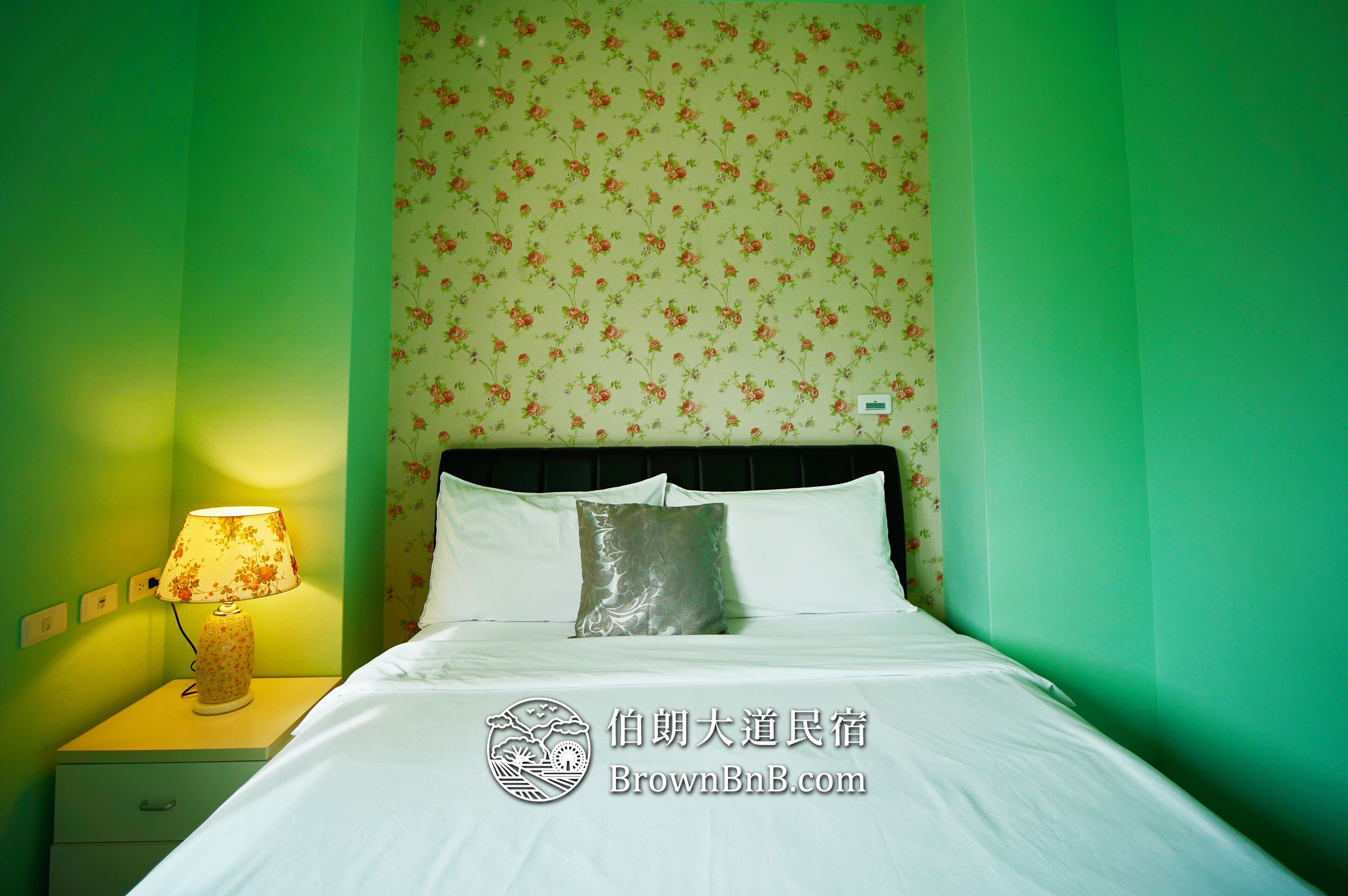 A neatly made bed with white linens and a single decorative pillow, a bedside lamp on a nightstand, and a floral wallpaper pattern on the wall.