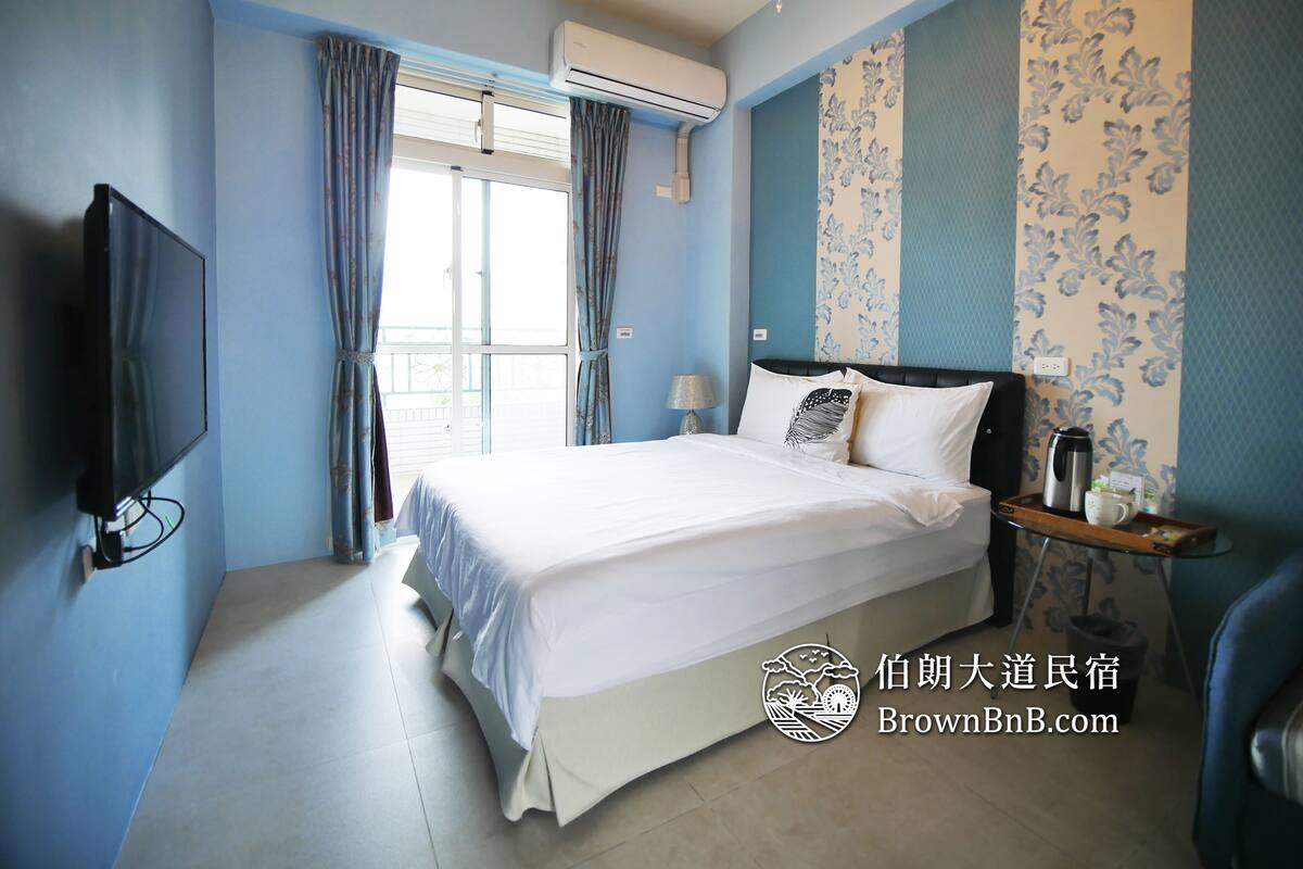 A hotel room with a large bed, a flat-screen TV, a bedside table with a lamp, and a window with curtains.